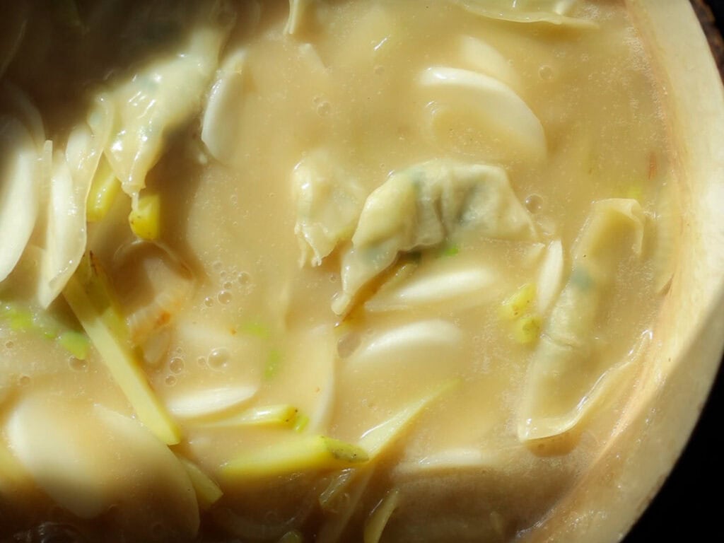 A close-up of a creamy vegan rice cake soup with dumplings and sliced vegetables. The mixture appears thick, with visible pieces of dumplings and green onion slices, all enveloped in a smooth, light-colored broth.